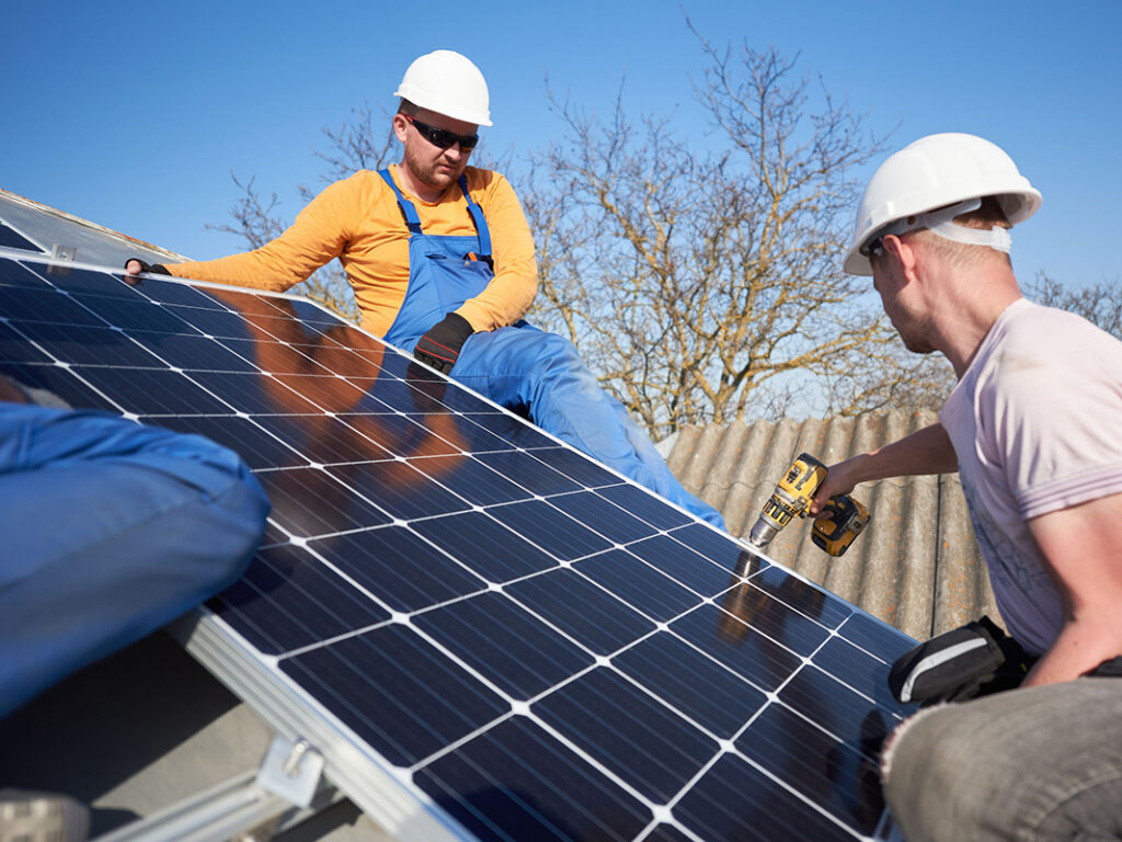 Male team workers installing stand-alone solar photovoltaic panel system using screwdriver. Group of electricians mounting solar module on roof of modern house. Alternative energy ecological concept.